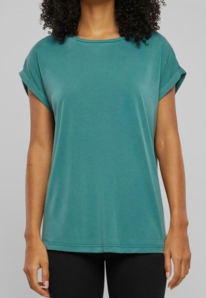 Frau mit lockigem Haar, die ein locker sitzendes, kurzärmeliges Shirt in Teal und schwarze Hose vor einem schlichten grauen Hintergrund trägt.