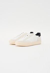 White sneakers with a suede and smooth leather upper, contrasting black heel, flat laces, and a textured cream rubber sole.