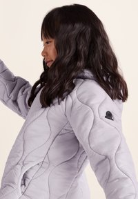 Light purple quilted jacket with a hood, featuring curved stitching patterns and a small logo on the left sleeve. Soft, lightweight material.