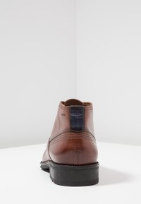 Botte chelsea en cuir marron avec tirette noire et semelle noire, vue de dos sur une surface blanche avec un fond blanc uni.