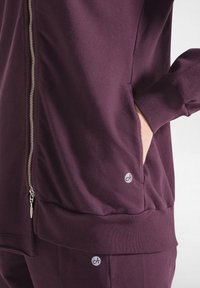 Giacca a zip viola realizzata in tessuto morbido, con tasche laterali e design aderente. Include un dettaglio con logo sull'orlo inferiore.