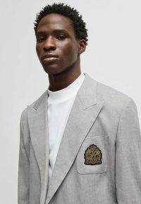 Light gray blazer with a textured fabric, featuring sharp lapels and a pocket patch with a gold emblem. Worn over a white high-neck top.