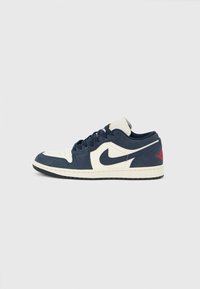 Nike Air Jordan 1 Low, άνω μέρος από μπλε σουέντ και κρεμ δέρμα, στρογγυλά κορδόνια, μαύρη σόλα, κόκκινα στοιχεία στη φτέρνα, διάτρητη περιοχή στα δάχτυλα, κλασική σιλουέτα.
