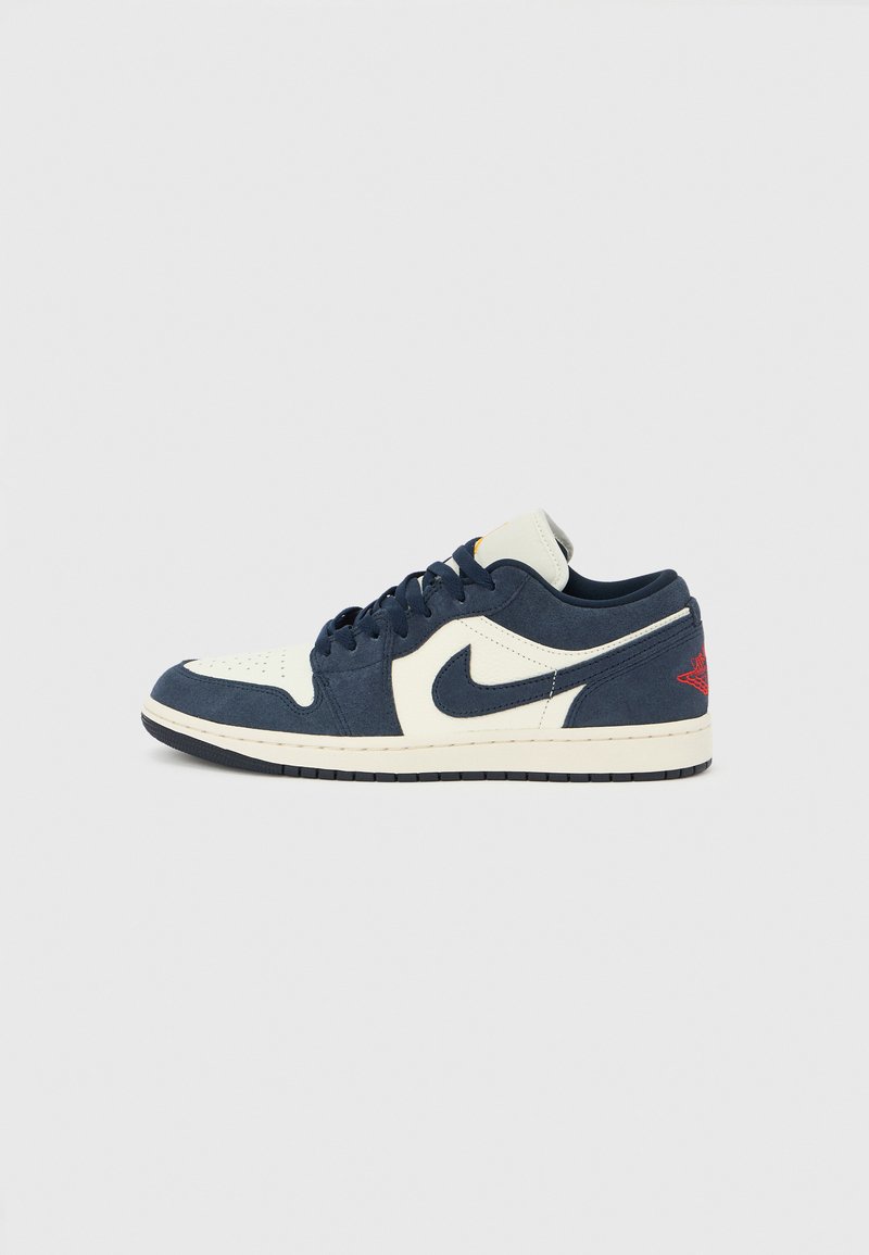 Nike Air Jordan 1 Low, άνω μέρος από μπλε σουέντ και κρεμ δέρμα, στρογγυλά κορδόνια, μαύρη σόλα, κόκκινα στοιχεία στη φτέρνα, διάτρητη περιοχή στα δάχτυλα, κλασική σιλουέτα.