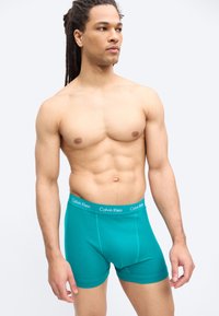 Μυώδης άντρας με κοτσιδάκια, φορώντας πετρόλ εσώρουχα Calvin Klein, κοιτάζει προς το πλάι μπροστά σε λευκό φόντο.