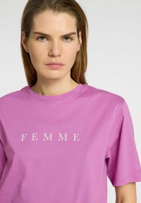Vaaleanpunainen lyhythihainen t-paita, joka on valmistettu puuvillasta ja jossa on pyöreä kaula-aukko; etuosassa on valkoisella painettuna sana "FEMME".