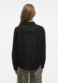 Melna, tekstūrēta krekla ar pūkainu, sapinušos virsmu, gariem piedurknēm un nedaudz pagarinātu aizmugurējo apakšmalu. Saskaņota ar leopardu raksta biksēm.