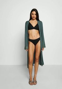 Femme portant un bikini noir, une longue robe ouverte teal avec des poignets en dentelle, et des chaussons à imprimé léopard, se tenant contre un mur lumineux et simple.