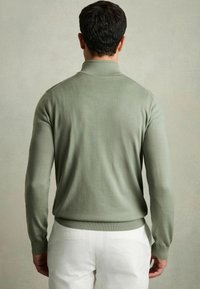 Pulover verde, cu mâneci lungi și guler, realizat dintr-un material neted și ușor. Dispune de manșete și tiv ondulate, purtat cu pantaloni albi.