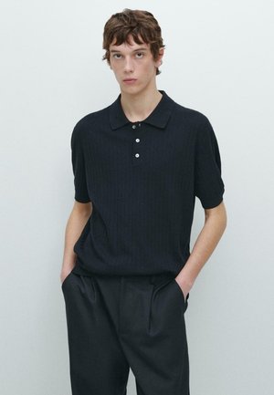 Ung mand med bølget brunt hår iført en sort kortærmet strikket poloshirt og sorte bukser, stående med hænderne i lommerne.