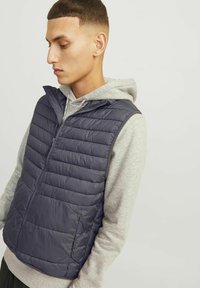Donkerblauwe puffer bodywarmer met horizontaal gestikt ontwerp, hoge kraag en rits, gedragen over een lichtgrijze hoodie.