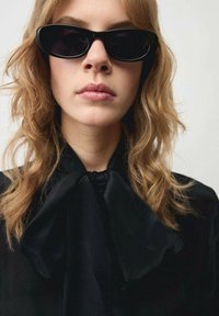 Zwarte cat-eye zonnebril wordt gedragen, aangevuld met een zwarte blouse met een grote strik bij de hals. Gelaagd haar valt op de schouders.