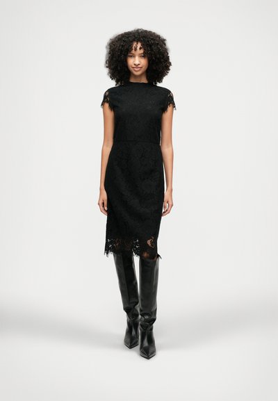 Femme aux cheveux bouclés portant une robe noire en dentelle à mi-longueur et des bottes en cuir noir montantes, se tenant contre un fond gris uni.