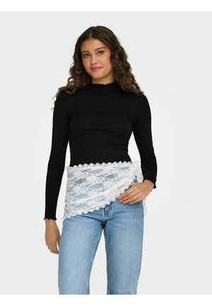 Woman with curly hair wearing a black long-sleeve top and light blue jeans, holding white lace fabric wrapped around her waist.