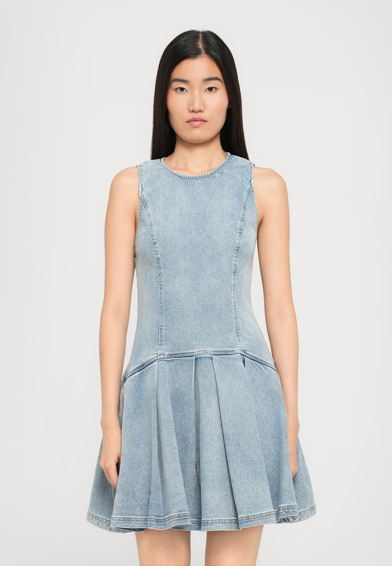 Young woman with long dark hair wearing a sleeveless, light blue denim dress with a fitted bodice and pleated skirt, standing against a plain background.