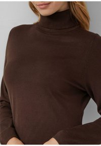 Kobieta ubrana w dopasowany, długi rękaw brązowy sweter z golfem, wykonany z miękkiego, gładkiego materiału.