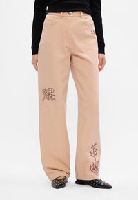 Μπεζ παντελόνια με φαρδύ πόδι και καφέ κεντημένα λουλούδια, φορεμένα με μαύρη μπλούζα και μαύρες μυτερές γόβες με ατσάλινα μάτια.