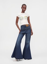 Femme portant un t-shirt court blanc avec des motifs de globe terrestre et un jean bleu taille haute à jambes larges, debout devant un fond uni.
