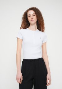 Λευκή μπλούζα με κοντά μανίκια, στρογγυλή λαιμόκοψη και πατιλέτα με κουμπιά, με ένα μικρό πράσινο λογότυπο στο αριστερό στήθος. Φορεμένη με μαύρο παντελόνι φόρμας.