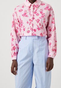 Blusa cropped floreale rosa con maniche lunghe, chiusura con bottoni e tessuto testurizzato. Abbinata a pantaloni a vita alta a righe blu chiaro.