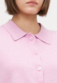 Maglione lavorato a maglia di un rosa chiaro con colletto, cinque bottoni rosa e una texture lucida, caratterizzato da una lieve brillantezza sparsa su tutto il tessuto.