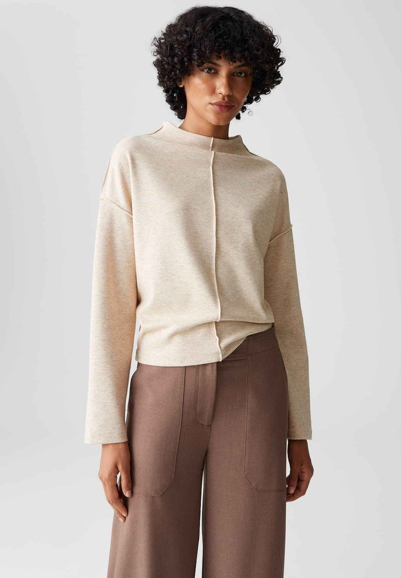Vrouw met krullend haar, gekleed in een beige lange mouwen top met een middennaad en hoog-geplaatste brede bruine broek tegen een effen achtergrond.