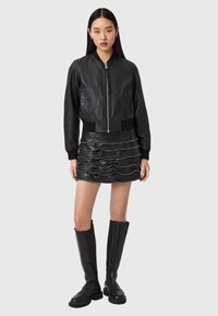 Γυναίκα που φοράει μαύρο δερμάτινο bomber μπουφάν, μαύρη μίνι φούστα με βολάν και μαύρες μπότες μέχρι το γόνατο, στέκεται μπροστά σε ανοιχτό γκρι φόντο.