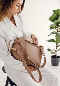 Hnědá strukturovaná kabelka s geometrickým designem, kožený materiál, zlaté kovové prvky a nastavitelné popruhy, kterou drží osoba v bílém oblečení.