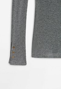 Chemise à manches longues côtelée grise avec trois boutons dorés près du poignet sur fond blanc.