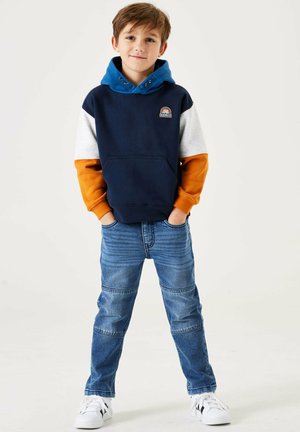 Felpa con cappuccio blu navy e blu con maniche grigie e arancioni, tasca frontale e toppa grafica; abbinata a jeans azzurri e sneakers bianche.