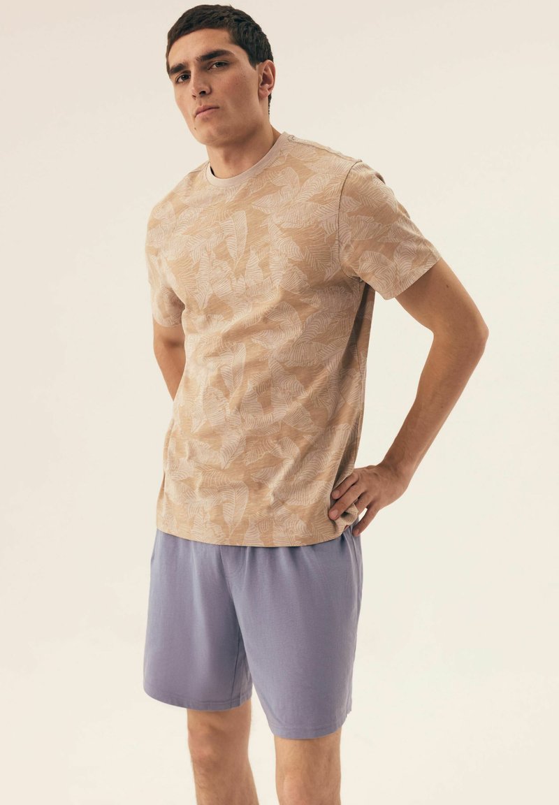 Beżowa koszula z krótkim rękawem w wzór liści, zestawiona z jasnopurpurowymi szortami. Gładka tekstura i luźny krój.