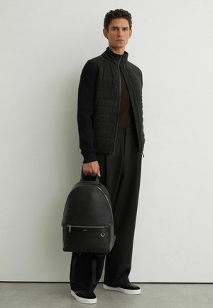 Man die een zwart jack en een zwarte broek draagt en een zwarte leren rugzak vasthoudt aan het handvat tegen een witte muur.