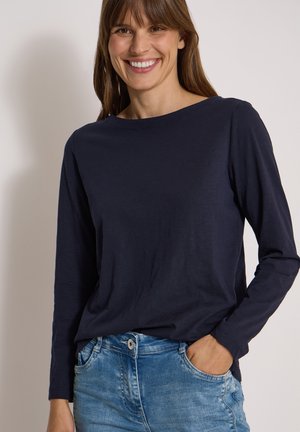 Femme souriante portant une chemise bleu marine à manches longues et un jean bleu, une main dans la poche, sur un fond uni.