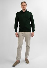 Man staat in een donkergroene kabelgebreide ritstrui, beige broek en lichtbruine instapschoenen tegen een effen lichte achtergrond.