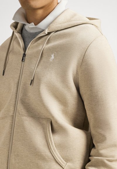 Μπεζ φούτερ με φερμουάρ, κατασκευασμένο από απαλό ύφασμα, με μπροστινή τσέπη, κουκούλα με κορδόνι και μικρό λευκό λογότυπο στο αριστερό στήθος.