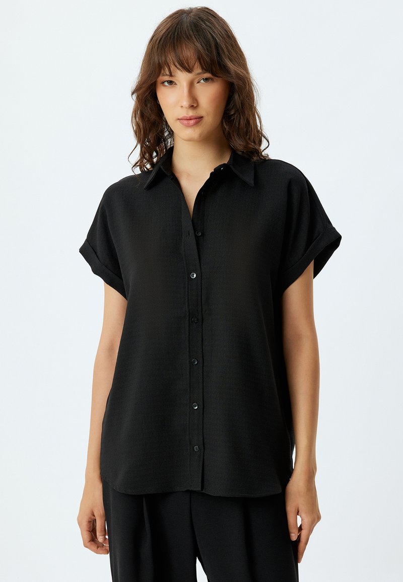 Camisa negra de manga corta y botones, confeccionada con una tela suave y texturizada. Cuenta con un cuello y puños enrollados, lo que ofrece una silueta relajada.