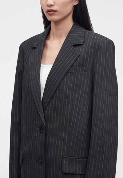 Femme portant un blazer sombre à fines rayures sur un haut blanc, vue de la poitrine vers le haut sur un fond uni.
