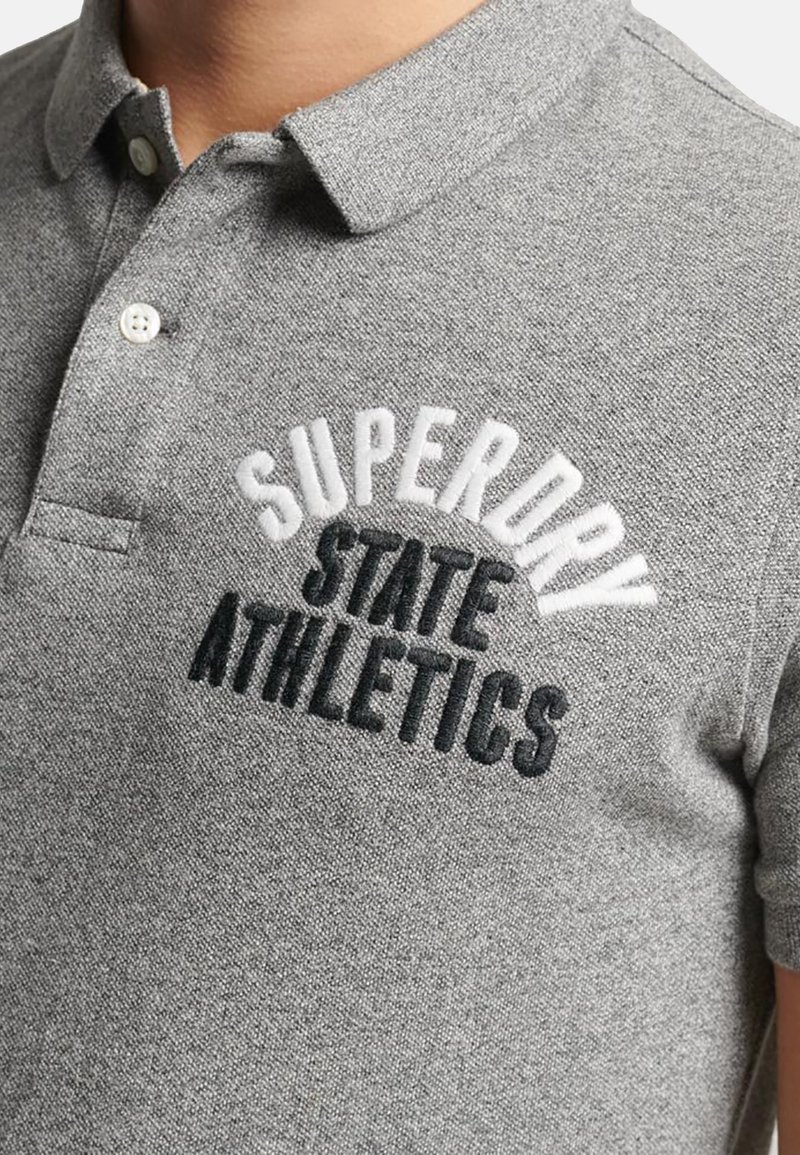 Camisa polo gris de tejido texturizado con un cierre de dos botones y logo bordado "Superdry State Athletics" en blanco y negro.