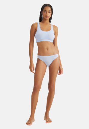 Kobieta z warkoczami, ubrana w jasnobluzy bezszwowy top sportowy i bikini, stojąca boso na tle jednolitego białego tła.