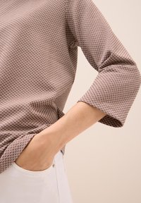 Hnědá košile s vzorem a tříčtvrtečními rukávy, vyrobená z texturované tkaniny, v kombinaci s bílými džíny. Viditelný výrazný geometrický design.