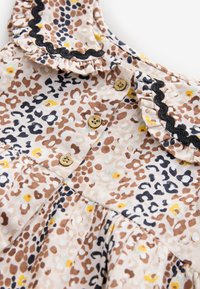 Leoparda raksta kleita bēšā krāsā ar brūniem, dzelteniem un melniem plankumiem, volāna apkakli un trim koka pogām priekšpusē.