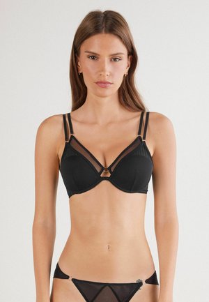 Woman wearing black lingerie set with sheer mesh panels and double shoulder straps, standing against plain white background.