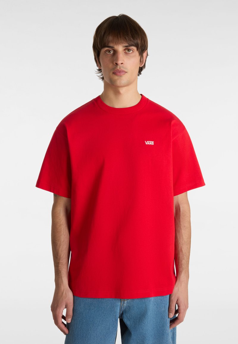 Camiseta de algodón roja con mangas cortas, cuello redondo y pequeño logo de Vans en el lado izquierdo del pecho. Llevada con jeans de mezclilla azul.