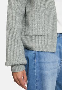 Gros plan sur une personne portant un pull en tricot gris clair avec des poignets et une poche avant, associé à un jean en denim bleu et un haut blanc.