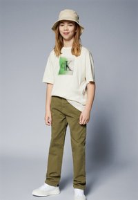 Ljusbeige t-shirt med grön grafik, i kombination med olivgröna byxor och vita sneakers. Hatten är också ljusbeige och fulländar outfiten.