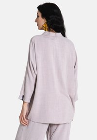 Blusa in mauve chiaro con vestibilità rilassata, maniche 3/4, colletto e tessuto strutturato. Indossata con pantaloni abbinati, che mostrano un design semplice.