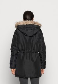 Parka noire avec capuche bordée de fourrure, taille cintrée, manches longues et poignets boutonnés. Fabriquée en matériau lisse et brillant, avec une coupe mi-longue.