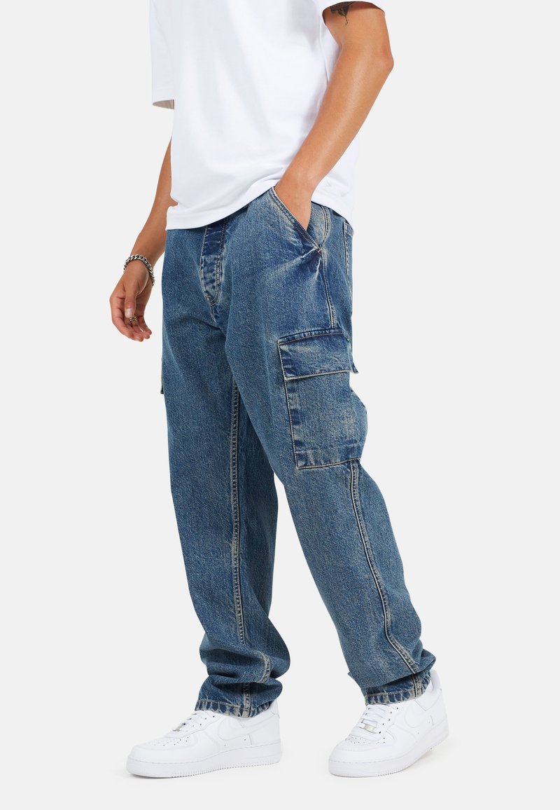 Pantaloni cargo in denim blu con tasche laterali, vestibilità comoda e dettagli di cucitura visibili, abbinati a sneakers bianche e una t-shirt bianca ampia.