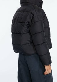 Zwarte pufferjas met een hoge kraag, gestikt ontwerp en een ritsluiting. Beschikt over dikke voering en een zijzak.