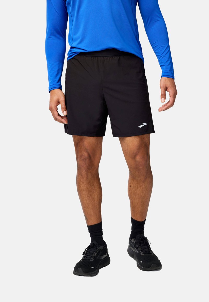 Pantalones cortos deportivos negros con una cinturilla elástica y un logo reflectante en la izquierda. Combinados con una camiseta de manga larga azul y zapatos negros.